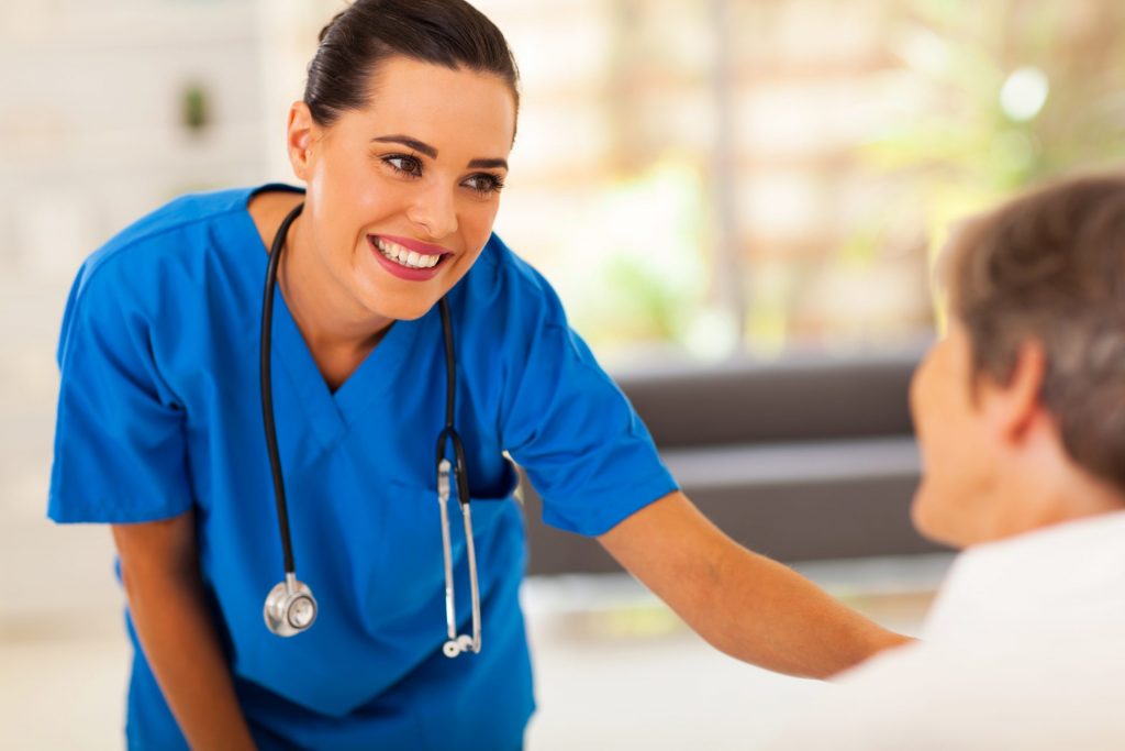 a female nurse in blue scrubs with a stethoscope around her neck looks towards an elderly woman