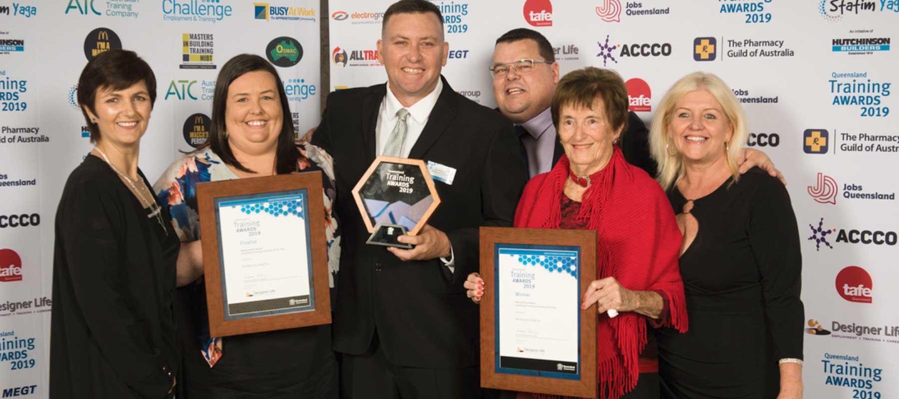 Six All About Living members formally dressed, taking a photo holding three Training Awards 2019 awards. Standing in front of a wall of brands including The Pharmacy Guild of Australia, Jobs Queensland, Tafe, MEGT, ACCCO