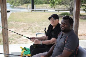 An elderly man and a carer sitting next to each other fishing and smiling, both men are wearing All About Living shirts