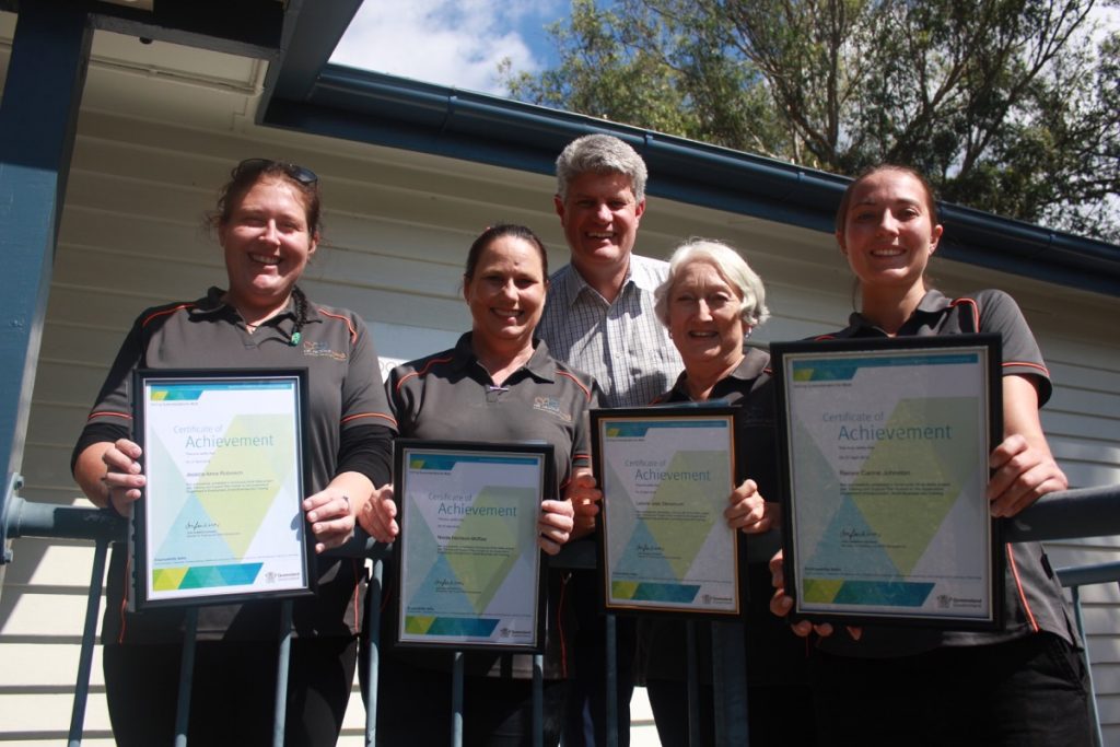 Four All About Living carers, nurses or volunteers smiling and holding certificate of achievement awards, a man in a grid suit stands behind them smiling