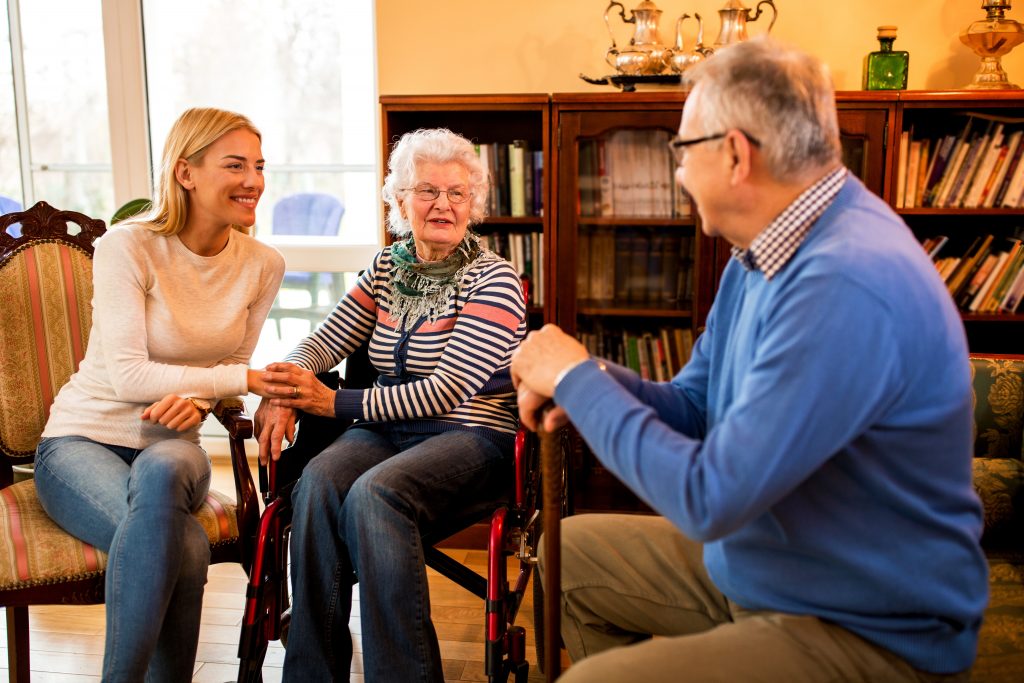 Sitting down, a woman holds the hand of an elderly woman in a wheel chair, who are both smiling towards an elderly man who is holding a walking stick and wearing a blue shirt