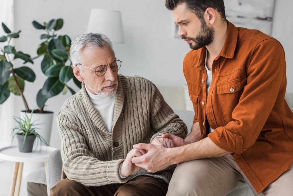 male home care nurse in orange shirt holding the hands of elderly man wearing a cardigan