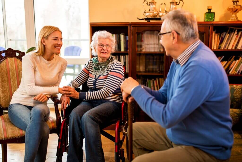 a woman holds the hand of an elderly woman in a wheel chair, who are both smiling at an elderly man