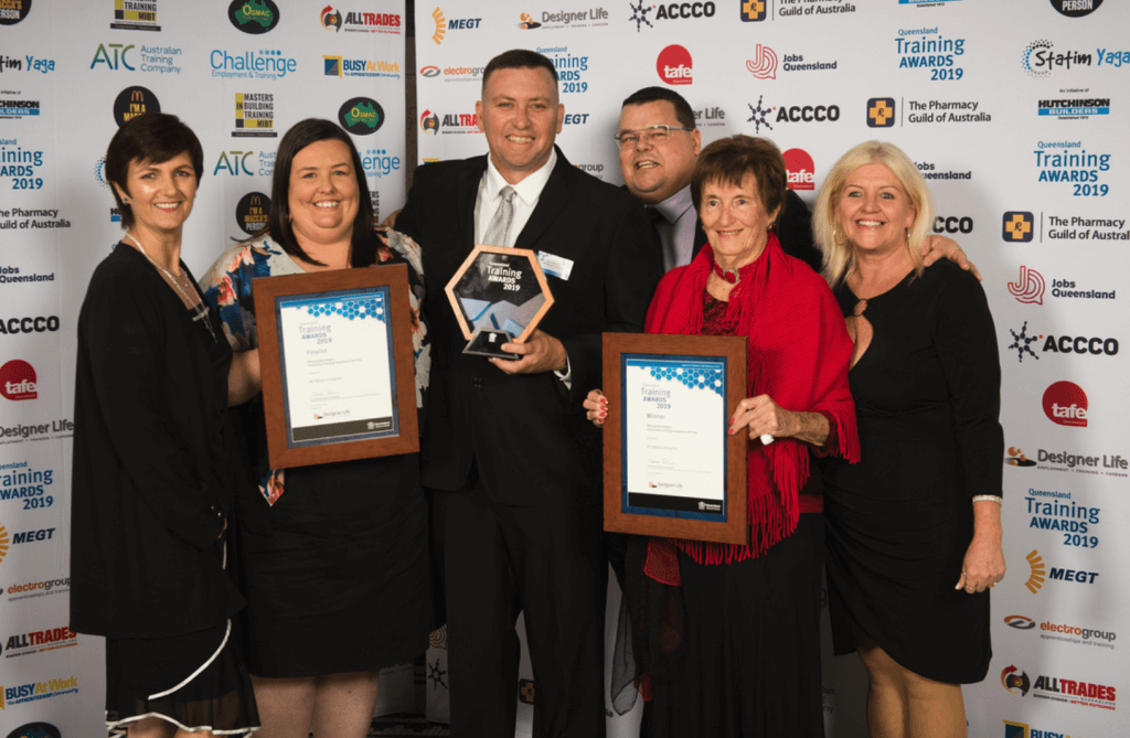 Six All About Living members formally dressed, taking a photo holding three Training Awards 2019 awards. Standing in front of a wall of brands including The Pharmacy Guild of Australia, Jobs Queensland, Tafe, MEGT, ACCCO